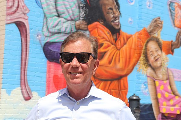 Governor Lamont with sunglasses standing in front of a mural.
