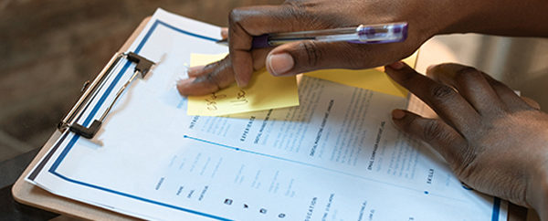 African American person reviewing and preparing a resume on a clipboard with a pen and sticky notes.