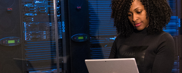 Young African American woman in black office clothing in front of a server room working on a laptop.